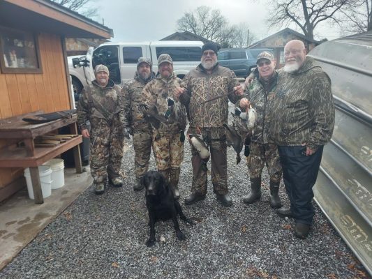 Six men in camouflage clothing stand together outdoors with a black dog, holding several ducks, next to a metal boat and a wooden building.
