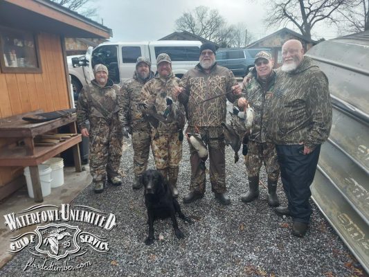 Six people in camouflage pose outdoors with ducks they have hunted, standing on gravel next to a boat and vehicle, with a black dog in front. "Waterfowl Unlimited Guide Service" logo is visible.