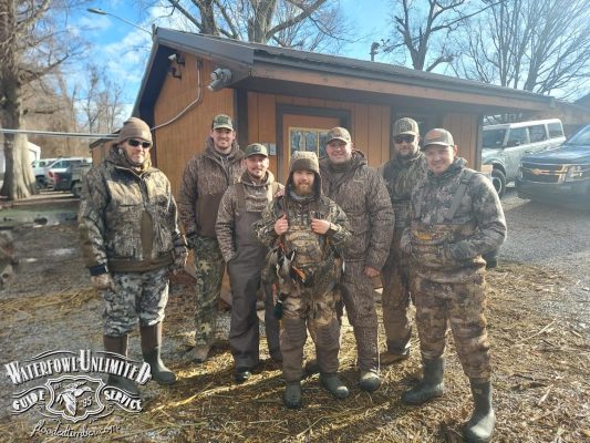 Seven people wearing camouflage outdoor gear stand together in front of a wooden building, with a "Waterfowl Unlimited Guide Service" logo in the lower left corner.
