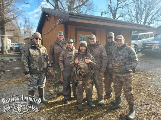 Seven people wearing camouflage outdoor gear stand together in front of a wooden building, with a "Waterfowl Unlimited Guide Service" logo in the lower left corner.
