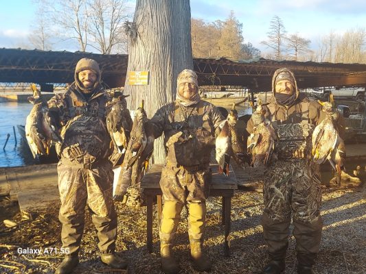 Three people in camouflage gear stand outdoors, each holding multiple ducks they hunted. There are trees and a dock in the background on a sunny day.