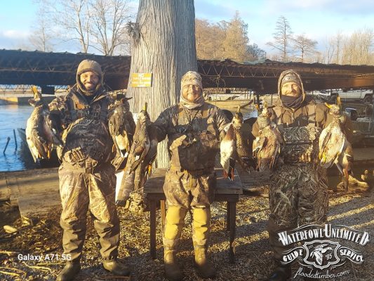 Three people in camouflage gear stand outdoors holding multiple ducks, with a dock, trees, and water in the background. A "Waterfowl Unlimited Guide Service" logo is visible in the corner.