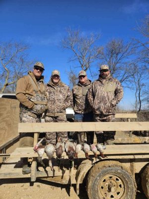 Four people in camouflage clothing stand on a trailer with several hunted birds displayed on the wooden rail in front of them; leafless trees and blue sky in the background.