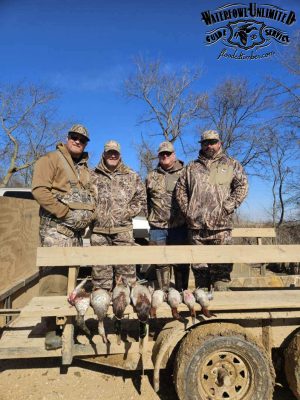 Four people in camouflage clothing stand behind a trailer with several hunted waterfowl displayed on it; leafless trees and a blue sky are in the background.