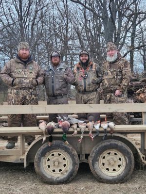 Four men in camouflage clothing stand behind a trailer with several hunted ducks laid out in front of them, with leafless trees in the background.
