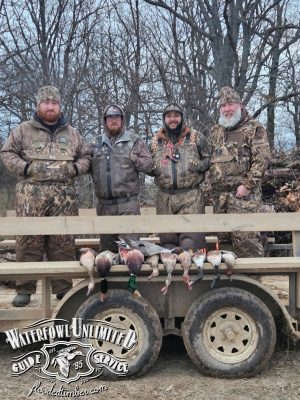 Four men in camouflage hunting gear stand behind a wooden rack displaying several ducks. The background shows leafless trees and a logo reads "Waterfowl Unlimited Guide Service.