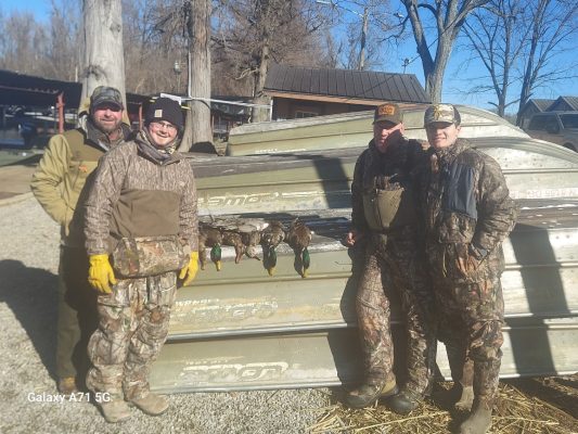 Four people in camouflage standing outdoors beside two metal boats with several ducks laid out on one boat. It is sunny and there are leafless trees in the background.