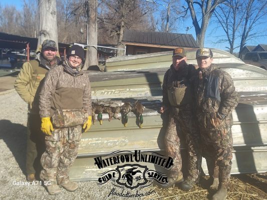 Four people in camouflage hunting gear stand beside a metal boat with several ducks lined up on it; "Waterfowl Unlimited" logo and website visible on the image.