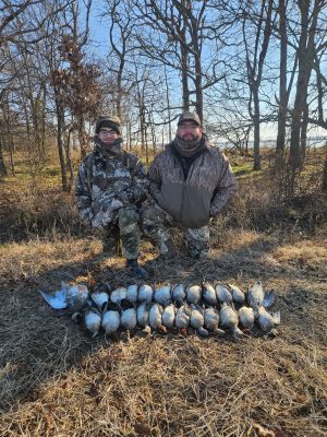 Two people in camouflage clothing kneel outdoors in a wooded area behind a row of harvested ducks laid out on the ground.