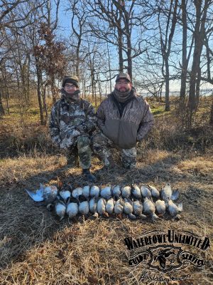 Two people in camouflage pose outdoors behind a row of dead ducks laid out on the ground, with leafless trees and dry grass in the background. The Waterfowl Unlimited logo is visible.