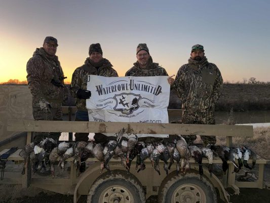 Four people in camouflage clothing stand behind a trailer displaying hunted ducks, holding a "Waterfowl Unlimited" banner outdoors at sunrise or sunset.