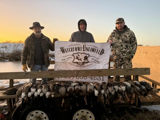 Three men stand behind a "Waterfowl Unlimited" banner, posed outdoors at sunrise with a display of hunted ducks arranged on a railing in front of them.