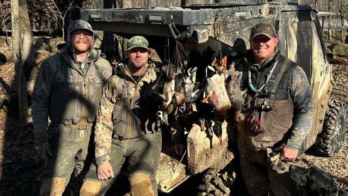 Three men in camouflage hunting gear stand by a muddy ATV, displaying several ducks they have hunted, with a forested background.