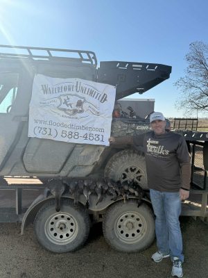 A man stands next to an ATV with a "Waterfowl Unlimited" banner and harvested ducks lined up on a trailer.