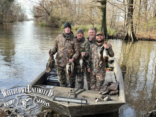 Four people in camouflage clothing stand on a boat holding ducks they have hunted, surrounded by trees and water. The boat features the logo "Waterfowl Unlimited Guide Service.