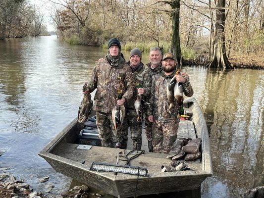 Four people in camouflage clothing stand on a small boat holding several ducks, with trees and water in the background.