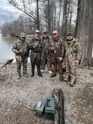 Five people in camouflage gear stand on a lakeshore holding ducks, with trees in the background and hunting equipment on the ground nearby.