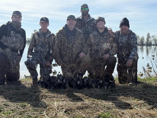 Six men in camouflage hunting gear pose by a lake with a collection of ducks arranged on the ground in front of them, with trees and water visible in the background.
