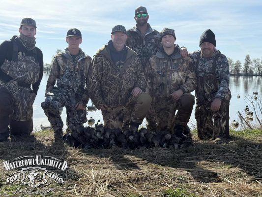 Six people in camouflage hunting gear pose by a lakeshore with several ducks on the ground in front of them. "Waterfowl Unlimited Guide Service" logo in the bottom left corner.