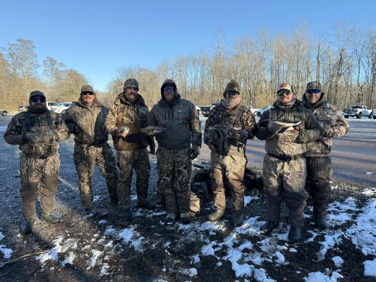 Seven people dressed in camouflage hunting gear stand on a snowy, gravel lot with bare trees and parked cars in the background, holding game birds.