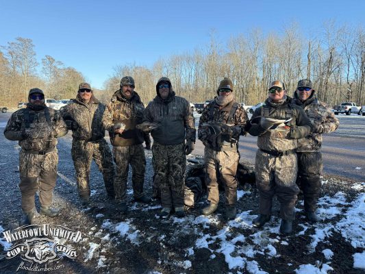 Seven people in camouflage clothing stand outdoors in a gravel lot holding hunted birds, with parked vehicles and leafless trees visible in the background.
