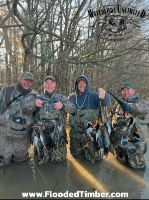 Four men in camouflage gear stand in shallow water holding several ducks, smiling at the camera. Text reads “Waterfowl Unlimited” and “www.FloodedTimber.com.”.