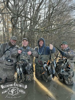 Four hunters in camouflage gear stand in shallow, wooded water, smiling and holding harvested ducks. "Waterfowl Unlimited Guide Service" logo is visible in the lower left corner.