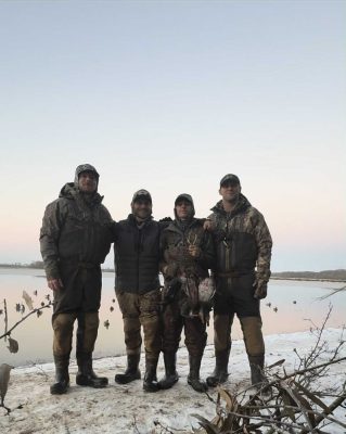 Four people dressed in camouflage and waders stand by a snowy lakeshore, holding several ducks, with a calm body of water and decoys in the background.