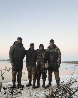 Four people in camouflage hunting gear stand arm-in-arm by a snowy lakeshore, holding ducks. The sky is clear, and decoys float on the water nearby.