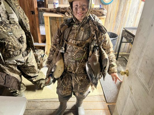 A person in camouflage hunting gear stands indoors with two ducks hanging from their chest harness, smiling at the camera.