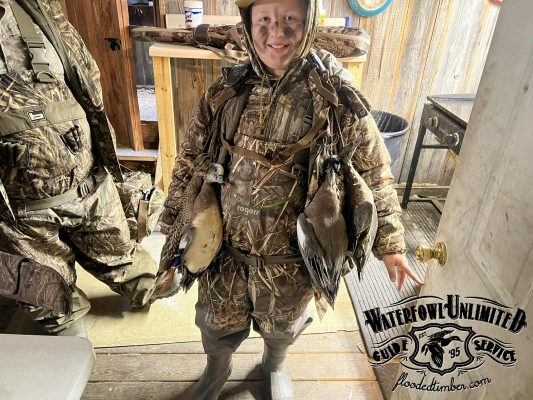 A person in camouflage hunting gear stands indoors holding several ducks, with "Waterfowl Unlimited Guide Service" logo visible in the corner.
