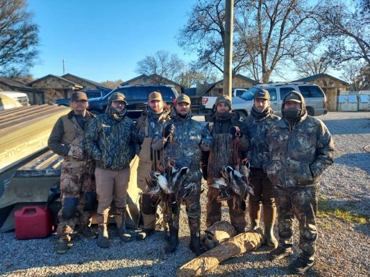 Six people in camouflage hunting gear stand outdoors holding harvested ducks, with vehicles and buildings in the background on a sunny day.