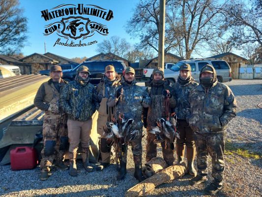 Six hunters in camouflage gear stand outdoors holding ducks in front of boats and cabins. A Waterfowl Unlimited logo appears at the top left of the image.
