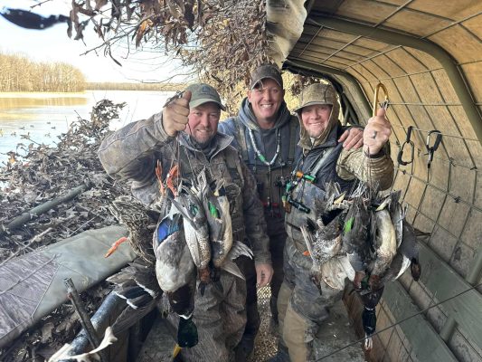 Three hunters in camouflage gear stand inside a duck blind, smiling and holding up several ducks they have hunted. The background shows a lake and leaf-covered surroundings.