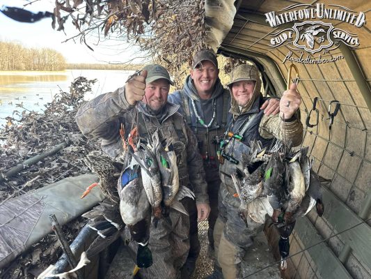 Three hunters in camouflage clothing pose in a duck blind, each holding several ducks. The logo for Waterfowl Unlimited Guide Service appears in the upper right corner.