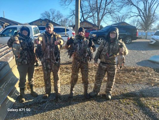 Four people dressed in camouflage gear stand outdoors holding up ducks they have hunted. Vehicles and buildings are visible in the background on a sunny day.