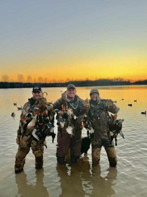 Three people in camouflage gear stand in shallow water at sunset, each holding several ducks they have hunted; decoy ducks float in the background.