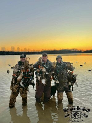 Three people in camouflage gear stand knee-deep in water at sunrise, each holding several ducks with decoys and water in the background. Logo in corner reads "Waterfowl Unlimited Guide Service.