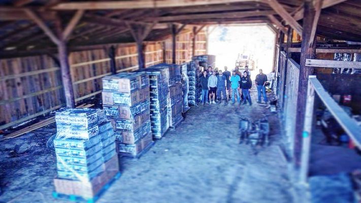 A group of people stands at the end of a barn-like structure with stacks of boxes organized in rows along the left side.