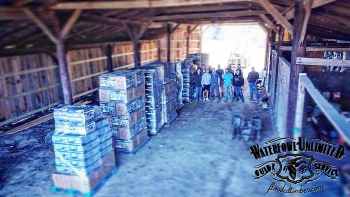 A group of people stands at the far end of a large wooden storage building filled with neatly stacked boxes; a "Waterfowl Unlimited Guide Service" logo is overlaid in the bottom right corner.