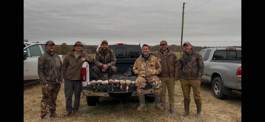 Six men in camouflage and outdoor gear stand and sit around a truck tailgate displaying several ducks, with vehicles and a field in the background under a cloudy sky.