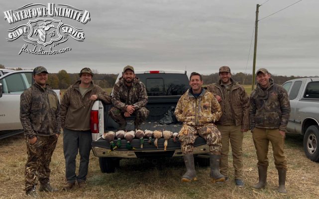 Six men in camouflage clothing stand and sit around a pickup truck, displaying several ducks on the open tailgate in an outdoor rural area.