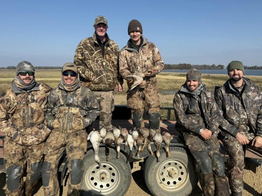 Six people in camouflage gear pose outdoors with a display of ducks laid out on a trailer in front of them, with fields and water in the background.