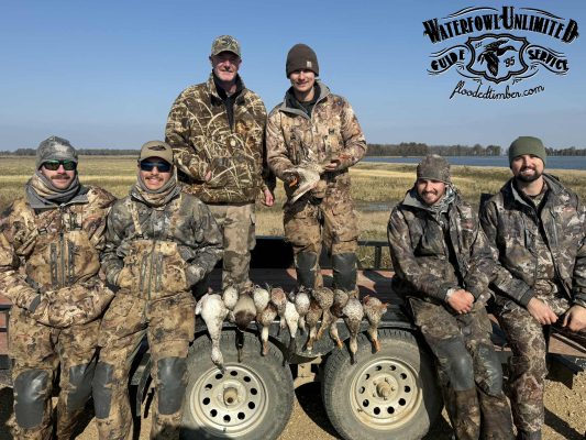 Six hunters in camouflage pose outdoors with a display of harvested ducks laid out in front of them. A "Waterfowl Unlimited" logo is visible in the upper right corner.
