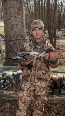 A person in camouflage gear holds a duck, with several other ducks lined up on a bench in front of a wooded background.