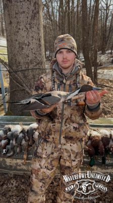 A person in camouflage hunting gear holds a duck with one hand; several ducks are lined up on a bench behind them. The logo "Waterfowl Unlimited Guide Service" is at the bottom right.